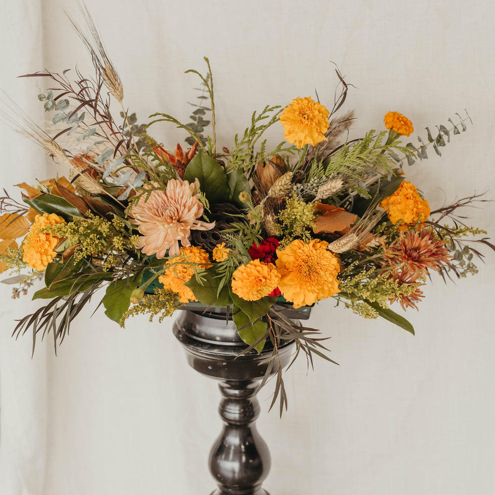 
                  
                    Floral arrangement with orange and green flowers on a dark pedestal against a light background
                  
                